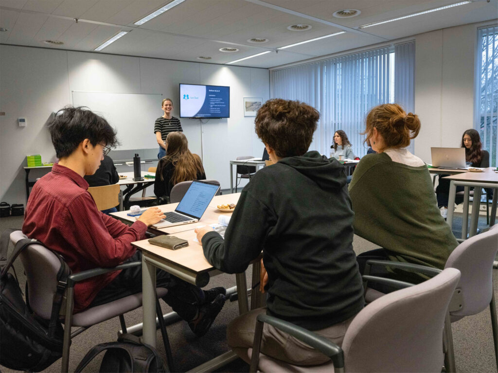 Een docent staat voor de klas tijdens een les Engels, met cursisten die aandachtig luisteren en meewerken.