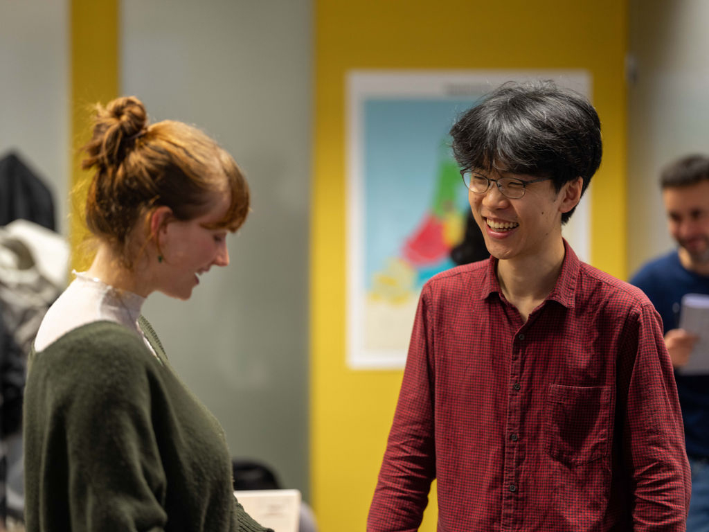Twee cursisten lachen terwijl ze in gesprek zijn tijdens een interactieve taalles, met een kleurrijke kaart op de achtergrond.