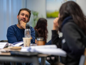 Cursist in een lesruimte kijkt nadenkend naar medecursist tijdens een gesprek aan tafel.