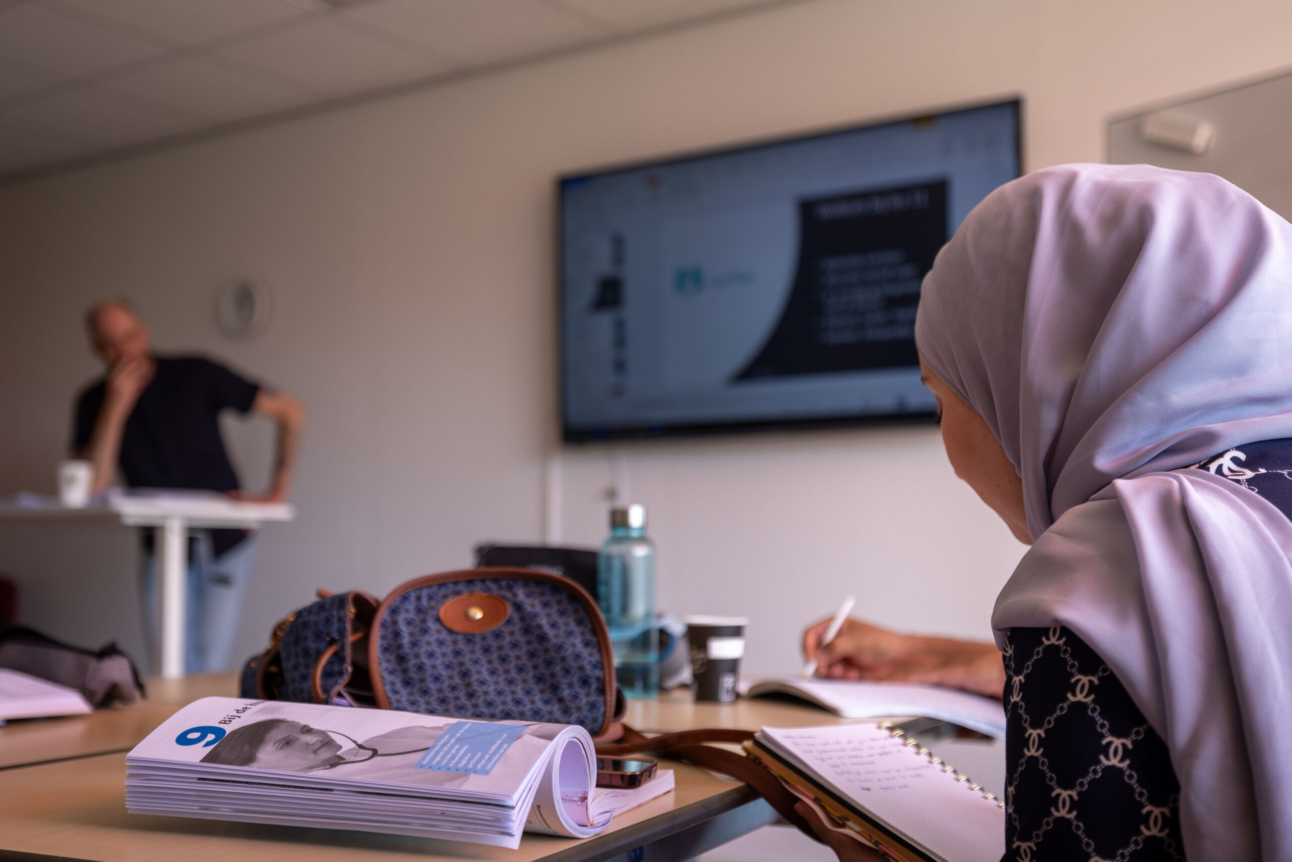 Een vrouwelijke cursist met hoofddoek schrijft in een schrift tijdens een taalles, terwijl een docent op de achtergrond lesgeeft bij een groot scherm.Een vrouwelijke cursist met hoofddoek schrijft in een schrift tijdens een taalles, terwijl een docent op de achtergrond lesgeeft bij een groot scherm.