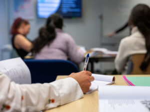 Close-up van een cursist die aantekeningen maakt tijdens de examentraining NT2, met op de achtergrond andere deelnemers in een klaslokaal.