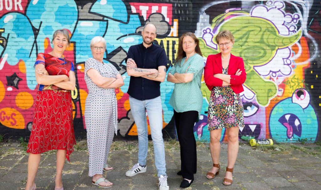 Teamleden van Lest Best poseren zelfverzekerd voor een kleurrijke graffitiwand in Rotterdam.